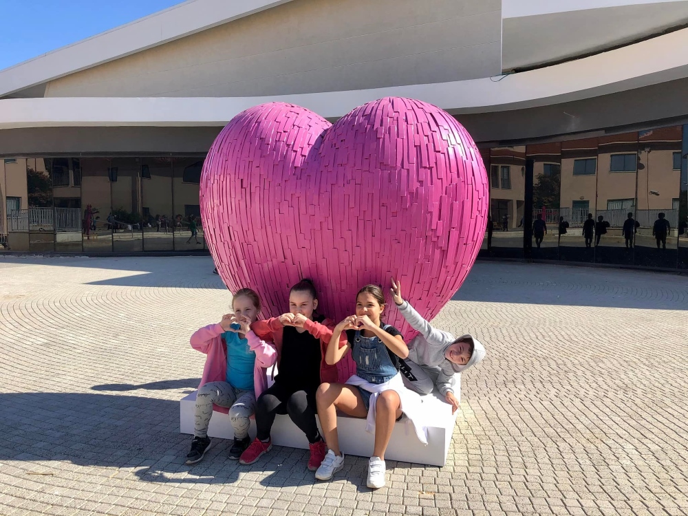 ילדות מסמנות בידיים לחיווי לב ויושבות לצד מיצב הלב הורוד במתחם המועצה האזורית דרום השרון